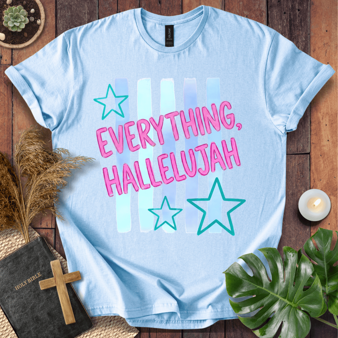 A light blue graphic t-shirt laid flat on a rustic wooden surface, surrounded by decorative props including a small plant, a candle, a book with a wooden cross, and a green leaf. The t-shirt features the text 'EVERYTHING, HALLELUJAH' in pink bubble letters with teal stars and pastel vertical stripes.