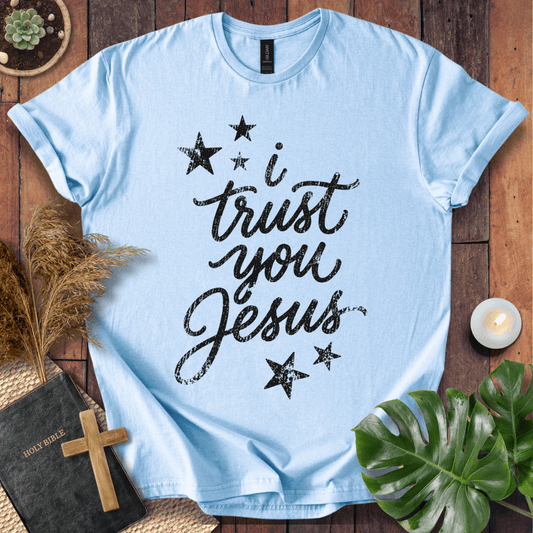 A light blue short-sleeve T-shirt laid flat on a rustic wooden surface, printed with the phrase 'i trust you Jesus' in black script and surrounded by black stars. The shirt is styled with a Bible, wooden cross, pampas grass, green leaves, and a small candle.