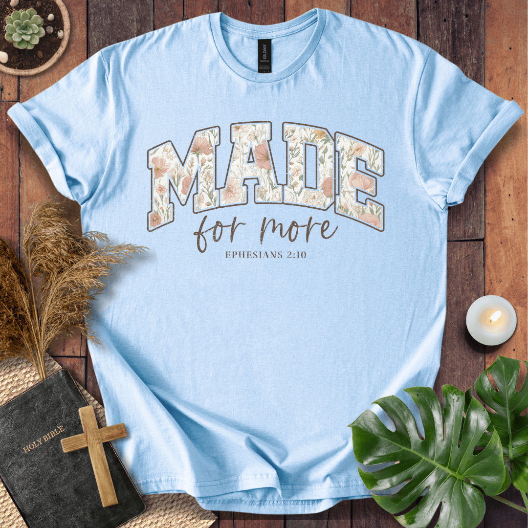 A light blue crewneck t-shirt laid flat on a rustic wooden surface, decorated with a Bible, a candle, pampas grass, a monstera leaf, and a small succulent. The shirt features a graphic that reads 'MADE for more' with 'EPHESIANS 2:10' underneath in brown script.