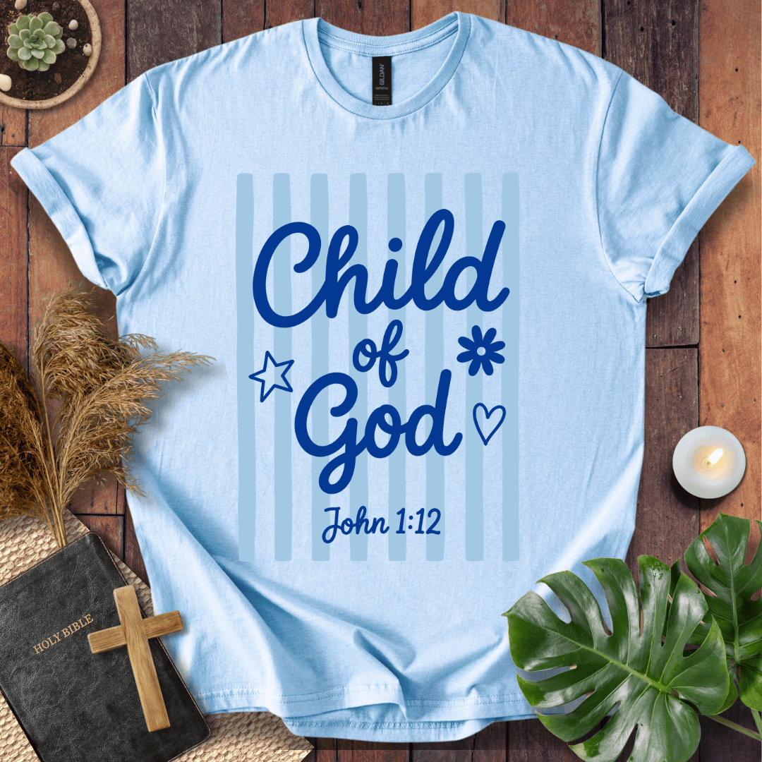 A light blue graphic t-shirt laid flat on a rustic wooden background, surrounded by decorative props including a Bible with a wooden cross, dried grass, a green monstera leaf, a small candle, and a succulent. The t-shirt features the text 'Child of God John 1:12' in dark blue script over a background of light blue vertical stripes.
