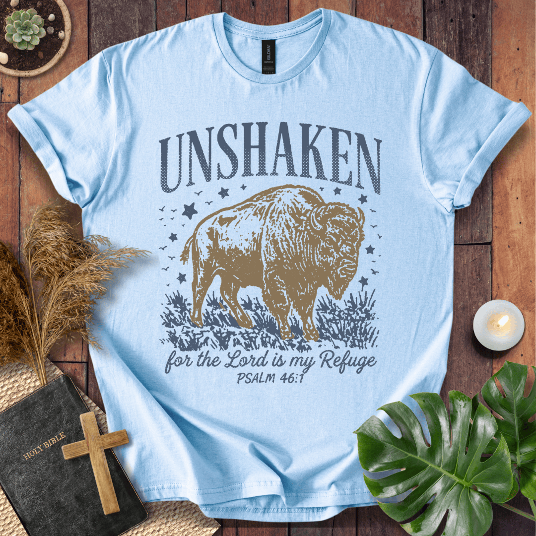 A light blue graphic t-shirt laid flat on a rustic wooden surface, surrounded by decorative props including a Bible with a wooden cross, pampas grass, a small candle, and green leaves. The t-shirt features a graphic of a bison with the text 'UNSHAKEN for the Lord is my Refuge PSALM 46:1'.