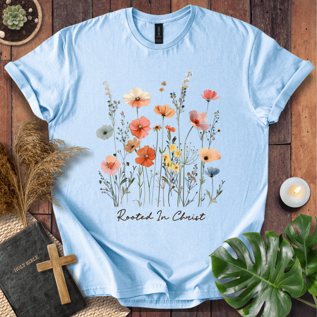A light blue short-sleeve t-shirt with a colorful watercolor floral print and the text 'Rooted In Christ' on the front, styled in a flat-lay arrangement on a wooden surface with a Bible, cross, pampas grass, a lit candle, and a green leaf.