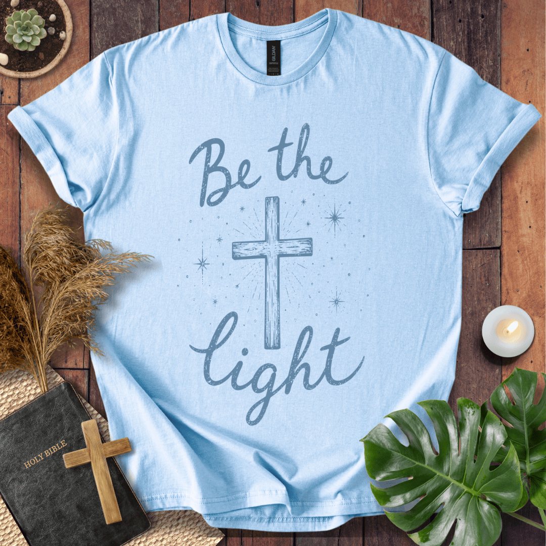 Christian t shirt for women with blue "Be the Light" text, a central cross, and starry accents on a wooden background.