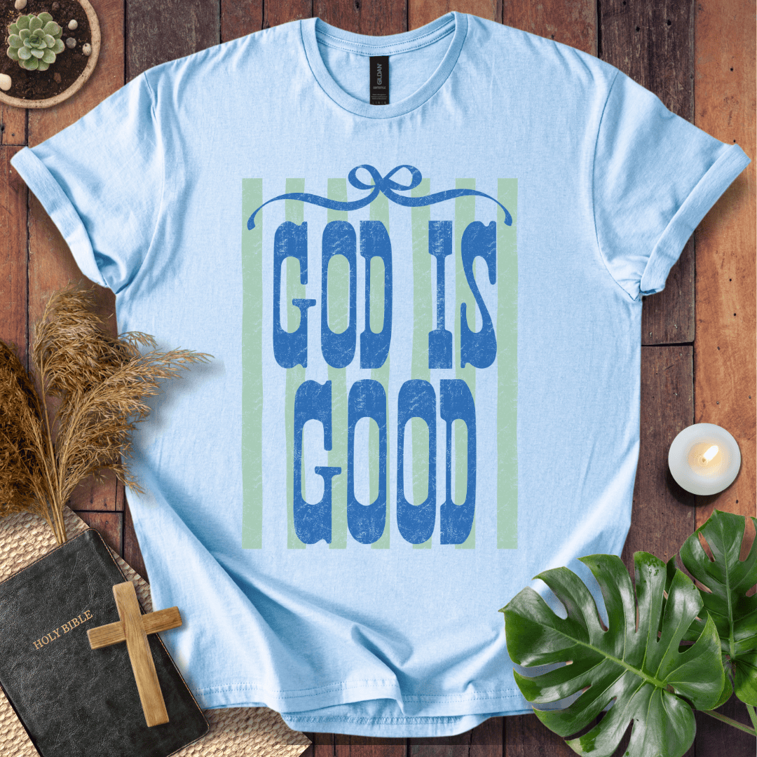 A light blue crewneck t-shirt with a graphic reading 'GOD IS GOOD' in textured blue letters over pale green vertical stripes, styled in a flat lay on a wooden surface with a Bible, cross, pampas grass, a candle, and a monstera leaf.