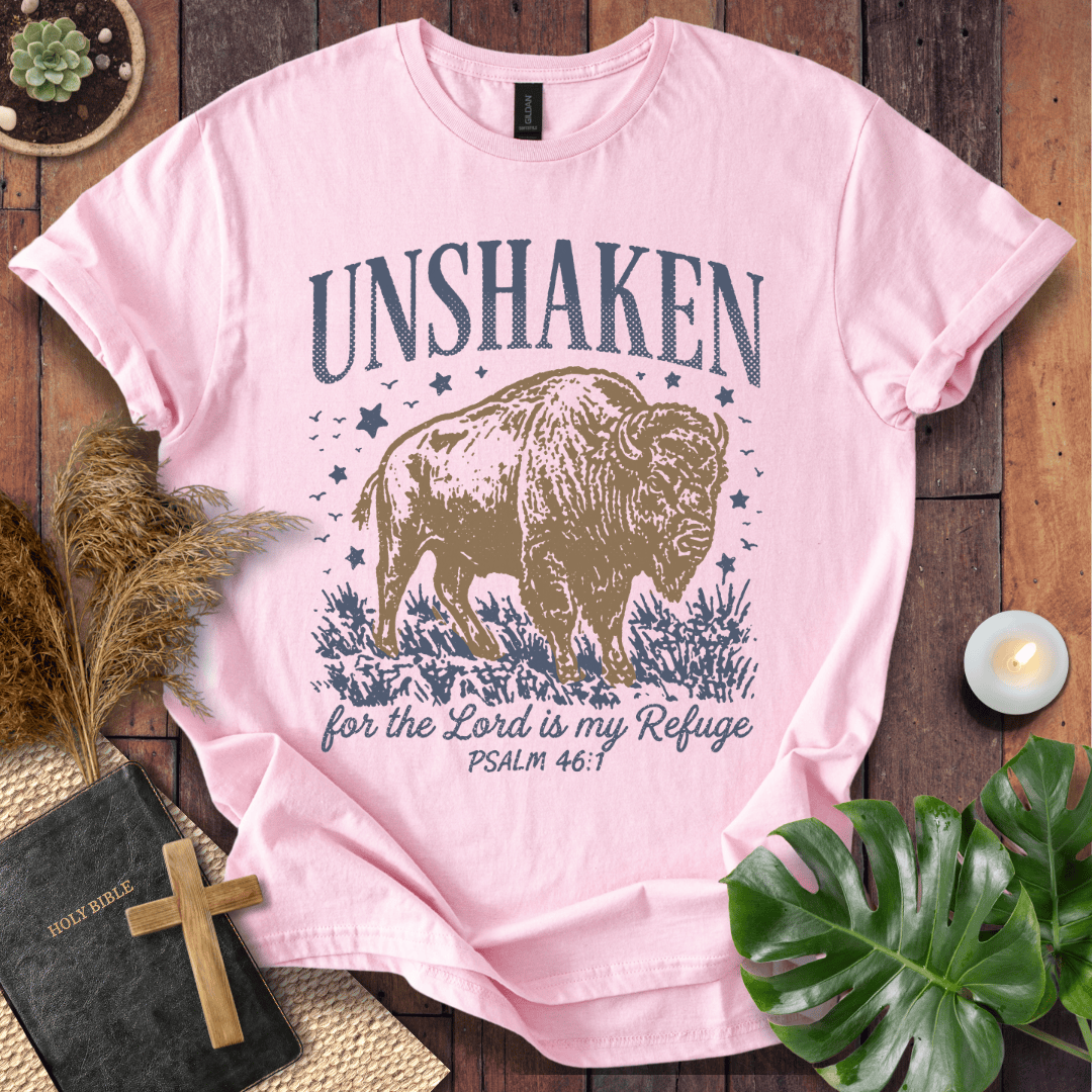 A light pink graphic t-shirt featuring a bison and the text 'UNSHAKEN for the Lord is my Refuge PSALM 46:1' in navy blue, styled in a flat-lay arrangement on a wooden background with a bible, cross, pampas grass, and green leaves.