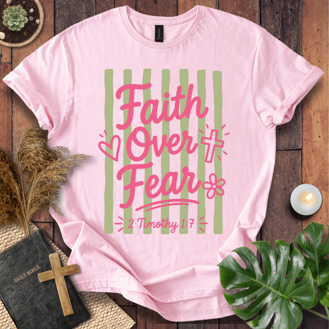 A light pink graphic t-shirt laid flat on a rustic dark wood background, surrounded by decorative props including a Bible, a wooden cross, dried grass, a green monstera leaf, and a small candle. The t-shirt features a graphic with vertical sage green stripes, pink text reading 'Faith Over Fear', and a cross icon with '2 Timothy 1:7' written below.