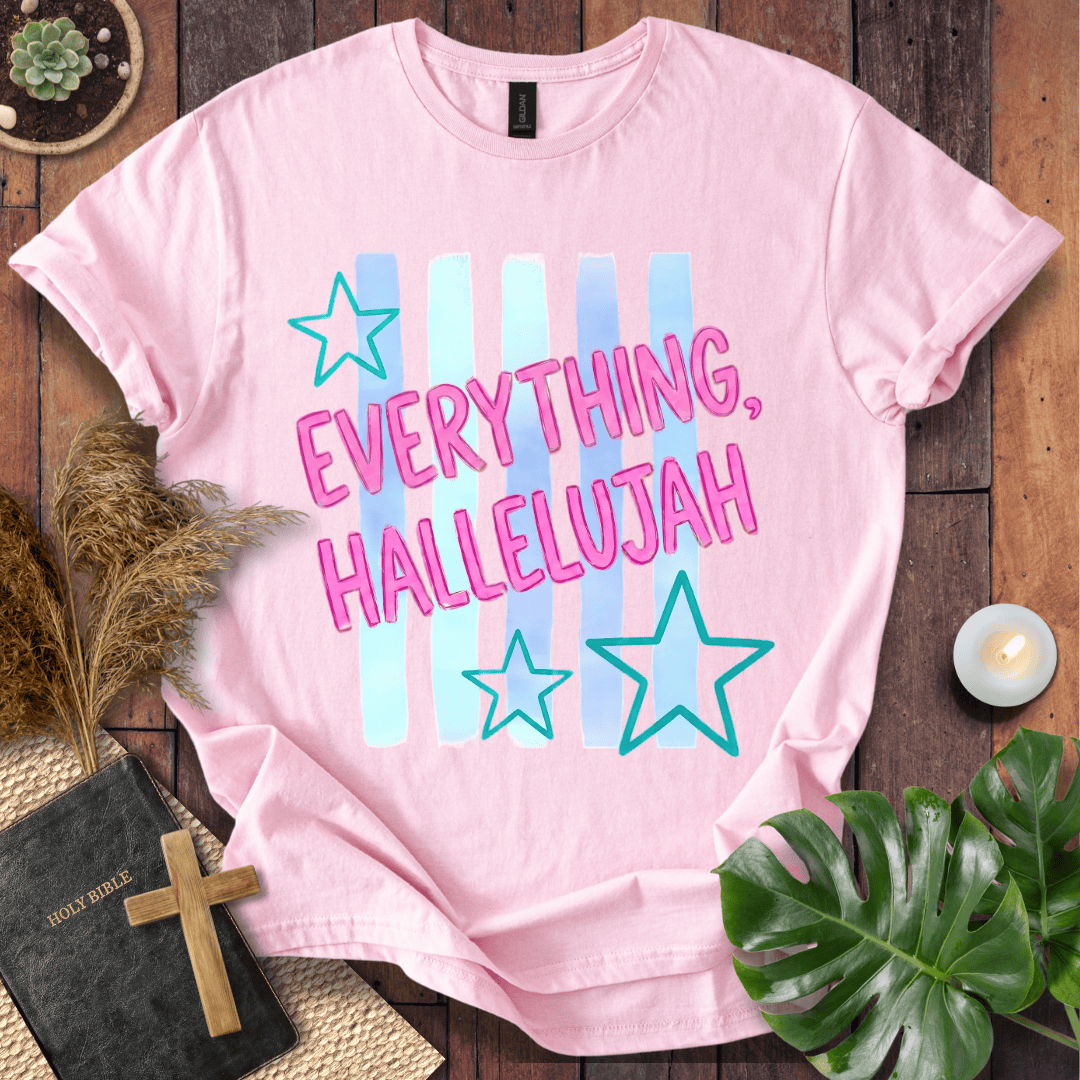 A light pink short-sleeve graphic T-shirt laid flat on a rustic wooden surface with decorative props including a Bible, cross, pampas grass, candle, succulent, and green leaves. The shirt has a pastel striped chest graphic with the phrase “EVERYTHING, HALLELUJAH” in pink letters and teal outlined stars.