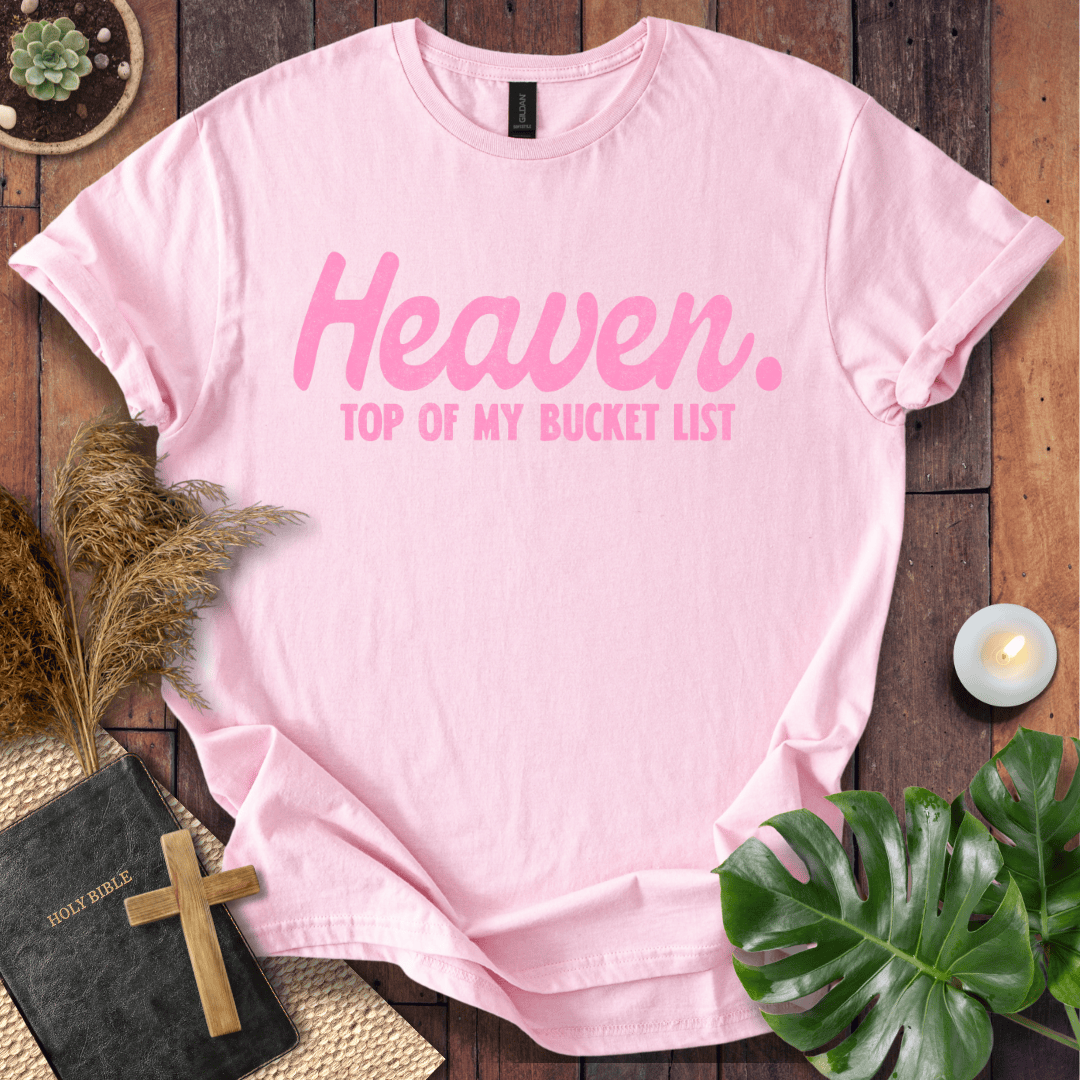 A light pink short-sleeve T-shirt laid flat on a rustic wooden surface, printed with the phrase 'Heaven. TOP OF MY BUCKET LIST' in pink lettering.