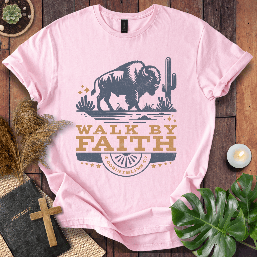 Pink t-shirt with a graphic of a bison and cactus, featuring the text 'Walk by Faith', on a wooden surface with decorative items.