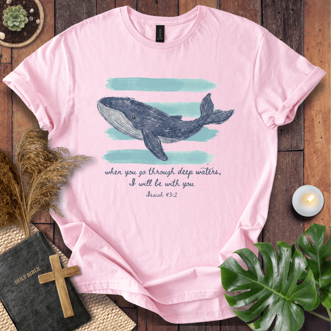 A light pink short-sleeve graphic T-shirt laid flat on a rustic wooden surface, featuring a whale illustration above the text, 'when you go through deep waters, I will be with you Isaiah 43:2.' Decorative props including a small candle, dried grass, a book with a cross, and green leaves surround the shirt.