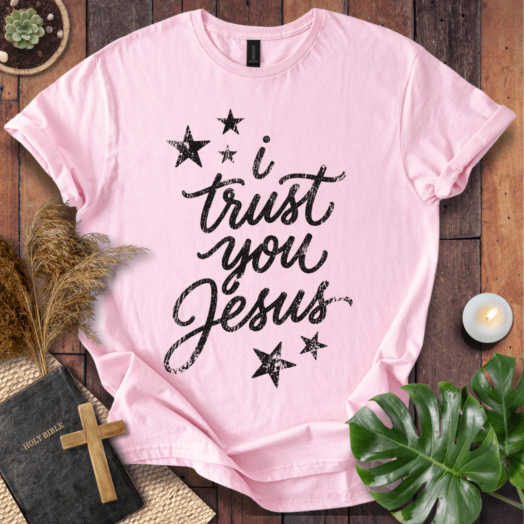 A light pink short-sleeve T-shirt laid flat on a rustic wooden surface, printed with the phrase 'i trust you Jesus' in black script and surrounded by black stars. The shirt is styled with a Bible, wooden cross, pampas grass, green leaves, and a small candle.