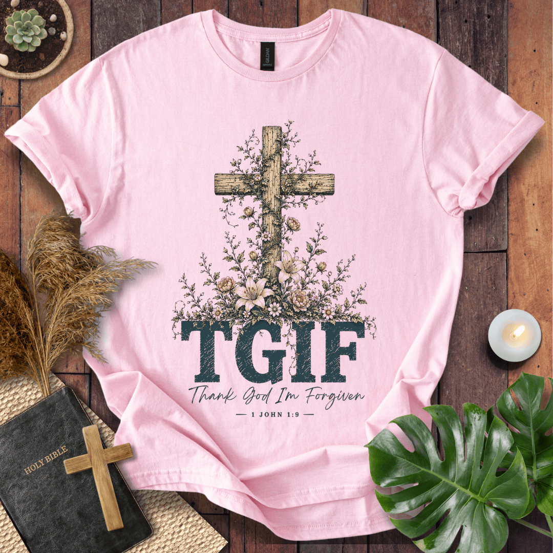 A light pink short-sleeve T-shirt laid flat on a rustic wood surface, featuring a large wooden cross surrounded by flowers and the text 'TGIF' with 'Thank God I'm Forgiven — John 1:9' underneath. Decorative props including a book, candle, dried grass, and green leaves surround the shirt.