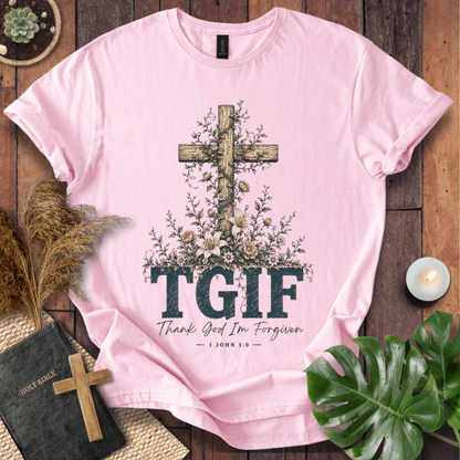 A light pink short-sleeve T-shirt laid flat on a rustic wood surface, featuring a large wooden cross surrounded by flowers and the text 'TGIF' with 'Thank God I'm Forgiven — John 1:9' underneath. Decorative props including a book, candle, dried grass, and green leaves surround the shirt.