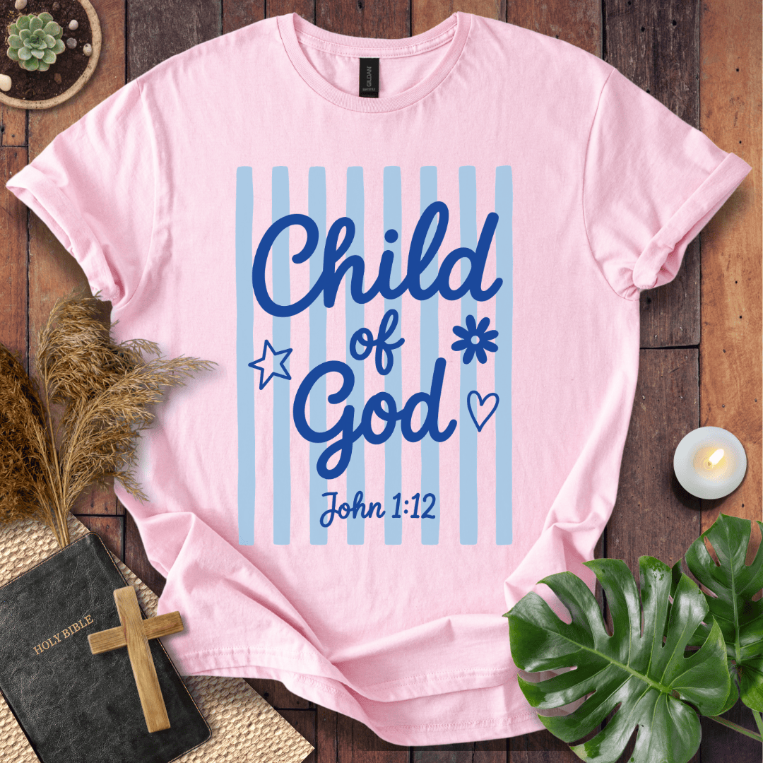 A light pink short-sleeve t-shirt with a blue graphic reading 'Child of God' and 'John 1:12' on a striped background, styled in a flat lay on a wooden surface with a Bible, cross, pampas grass, candle, and green leaves.