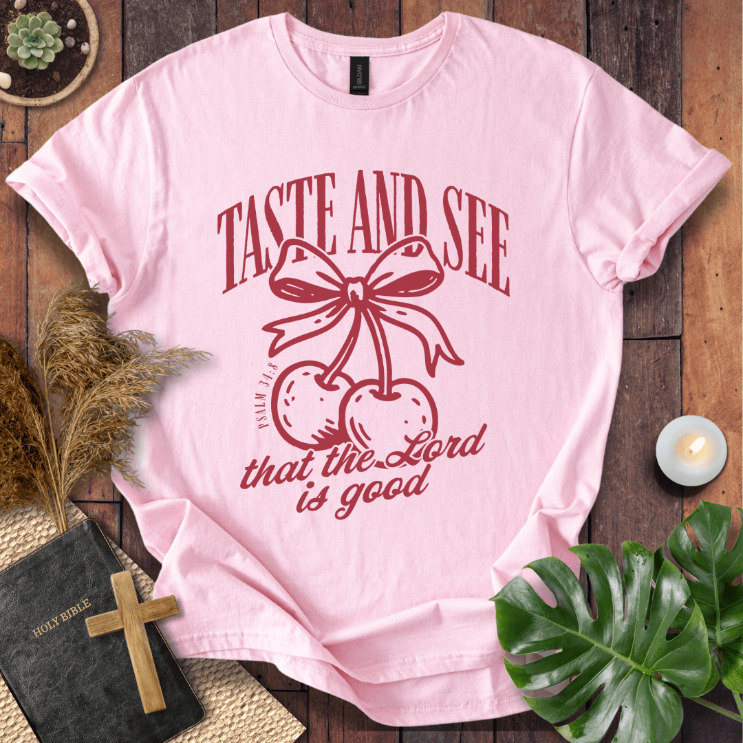 A light pink short-sleeve graphic T-shirt laid flat on a rustic wood surface, surrounded by decorative props including a Bible, cross, dried grass, green leaves, and a small candle. The shirt has a dark red printed design of cherries tied with a bow and the words 'TASTE AND SEE that the Lord is good.