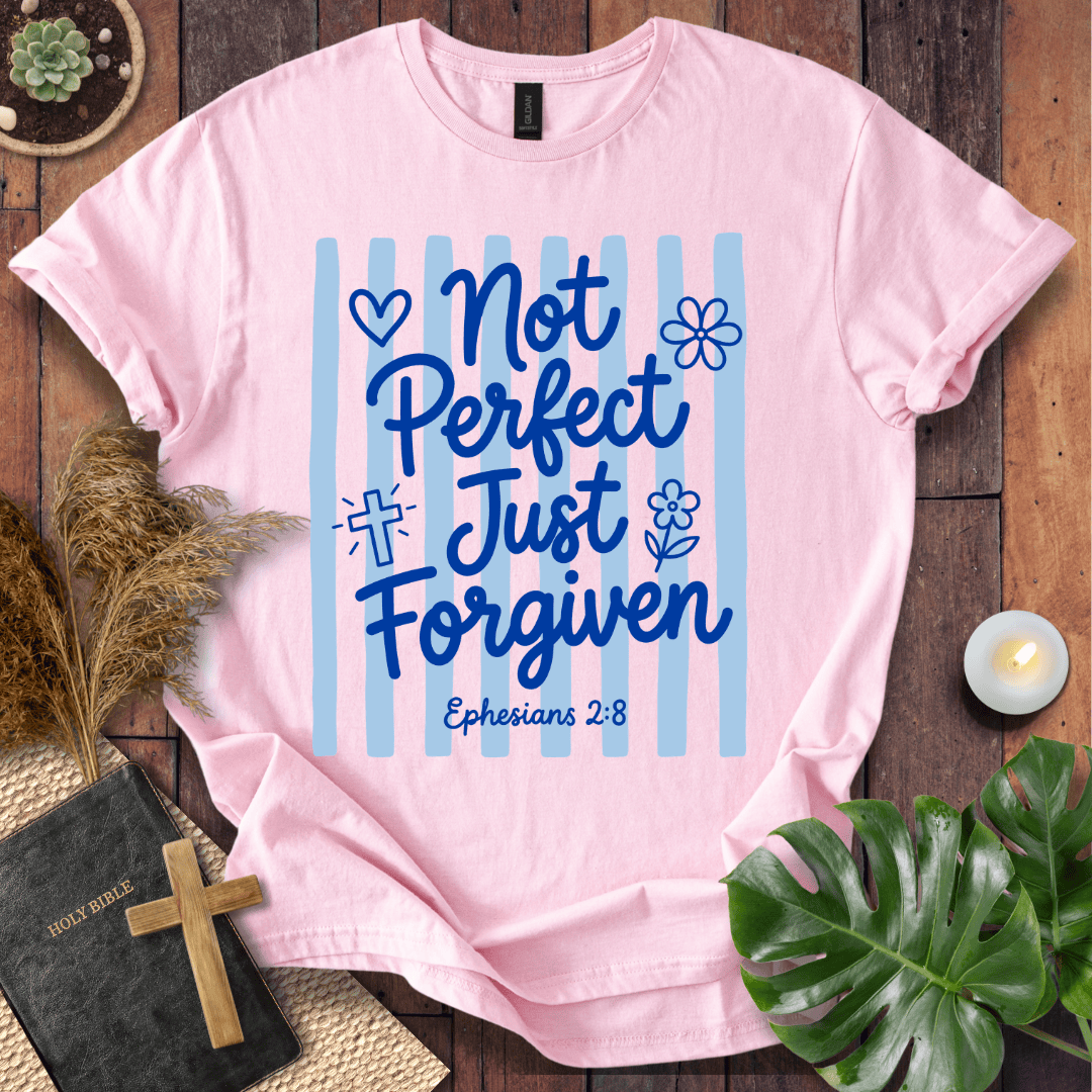 A light pink short-sleeve T-shirt laid flat on a rustic wooden surface, featuring the phrase 'Not Perfect Just Just Forgiven' with blue lettering, light blue vertical stripes, and 'Ephesians 2:8' printed below. The shirt is styled with a small book, wooden cross, dried grass, a candle, and green leaves around it.