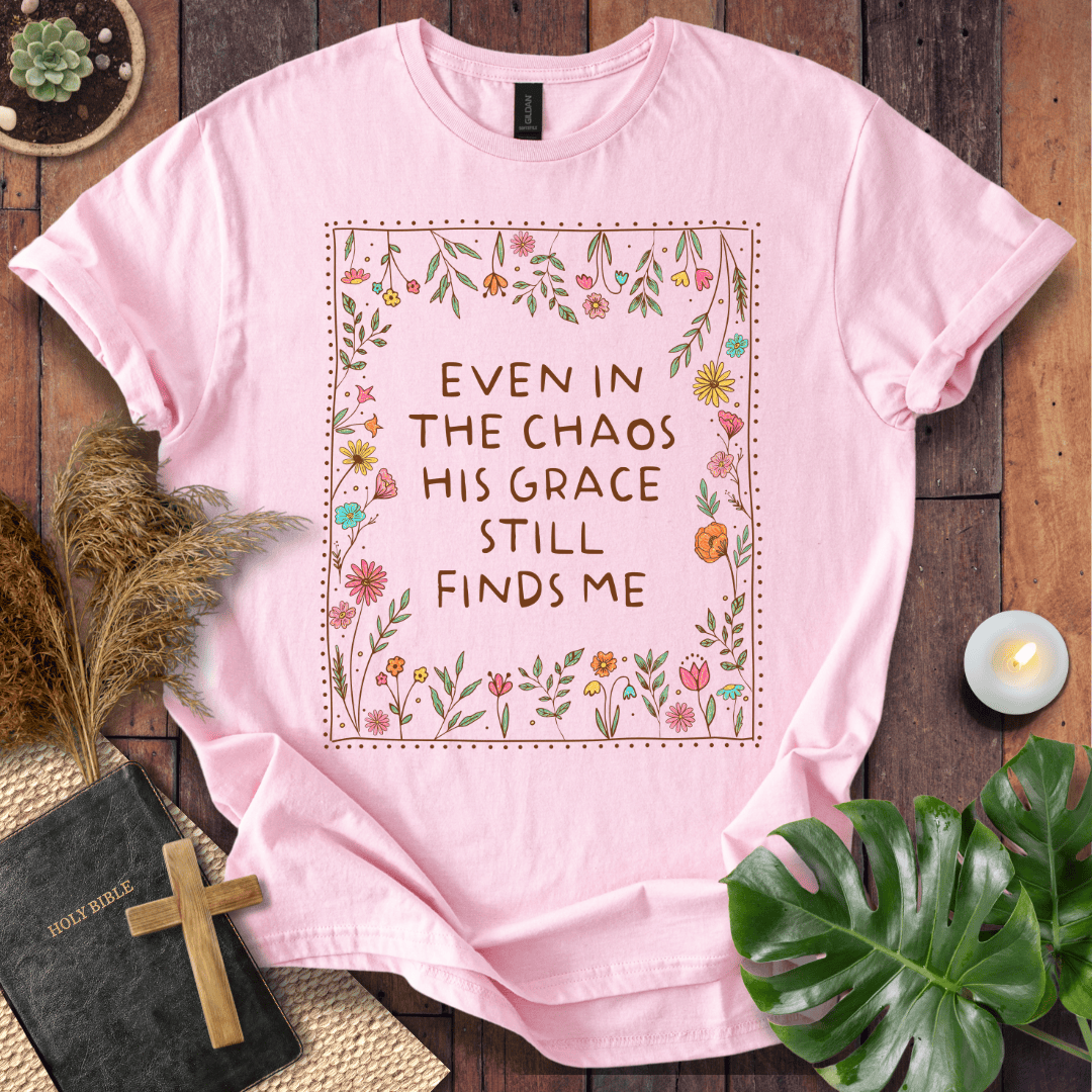 A light pink short-sleeve graphic T-shirt laid flat on a rustic dark wood surface, surrounded by decorative props including a small book with a wooden cross, dried grass, a lit candle, and green leaves. The shirt has a floral border and the phrase 'EVEN IN THE CHAOS HIS GRACE STILL FINDS ME' printed in brown text.
