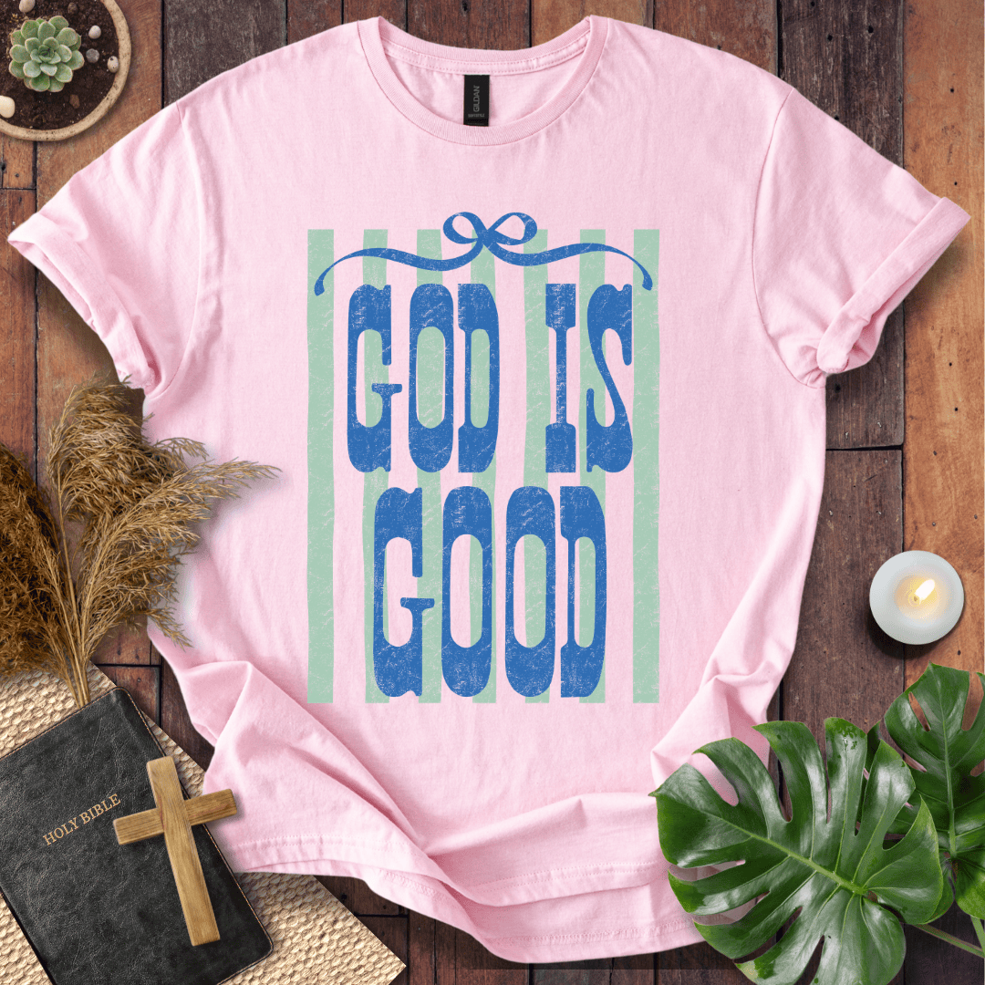 A light pink short-sleeve T-shirt laid flat on a rustic wooden surface, featuring a blue graphic that reads 'GOD IS GOOD' over pale green vertical stripes. The shirt is styled with a black Bible and cross, dried grass, a small candle, and green leaves.