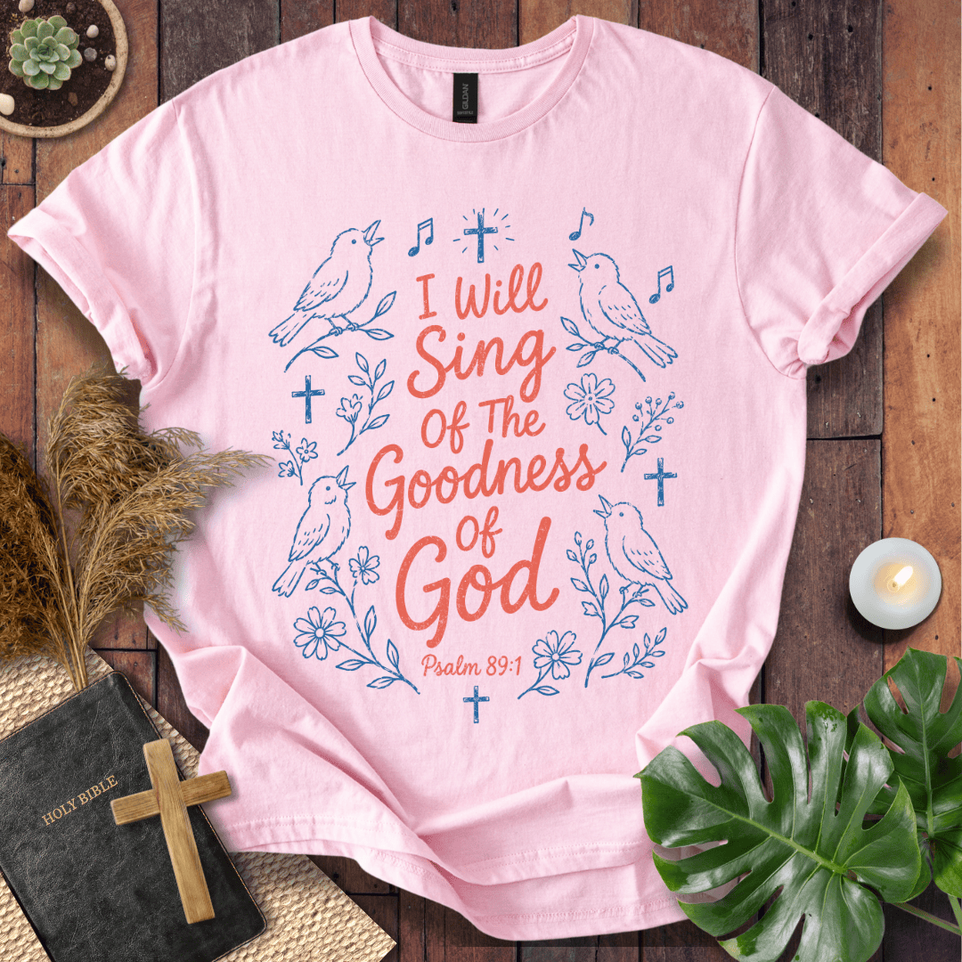 A light pink short-sleeve graphic T-shirt laid flat on a dark wood surface, surrounded by decorative props including a small plant, dried grass, a candle, a Bible with a wooden cross, and a green leaf. The shirt has a large front print with blue birds, flowers, music notes, and crosses, plus coral text reading 'I Will Sing Of The Goodness Of God' and 'Psalm 85:1'.