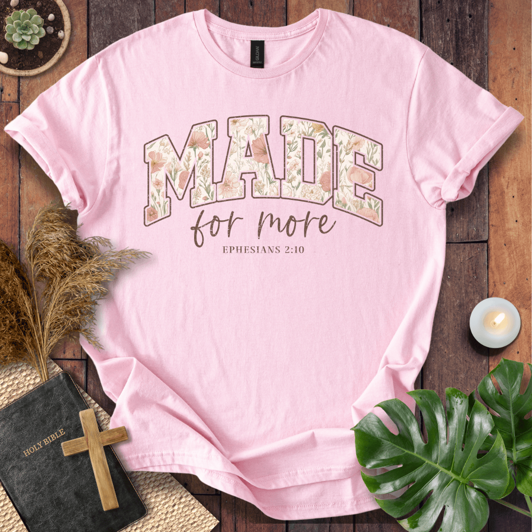 A light pink short-sleeve T-shirt laid flat on a rustic wooden surface, featuring the phrase 'MADE for more' with 'EPHESIANS 2:10' below. The word 'MADE' is filled with a floral pattern.