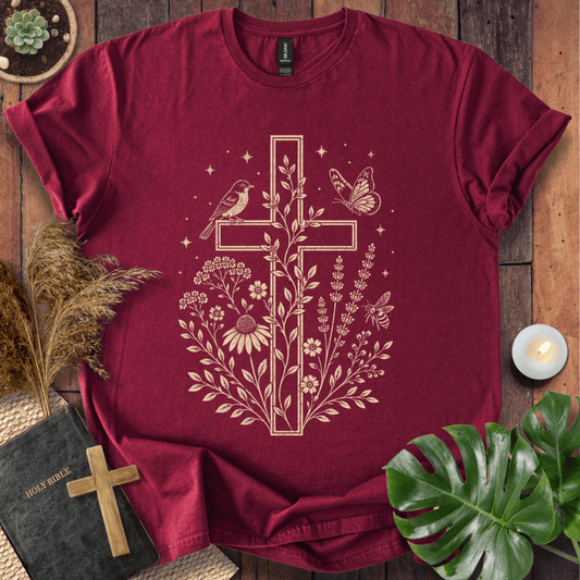 A dark red short-sleeve t-shirt with a beige cross graphic featuring birds, flowers, and butterflies, styled in a flat-lay arrangement on a wooden surface with a Bible, candle, and green leaves.