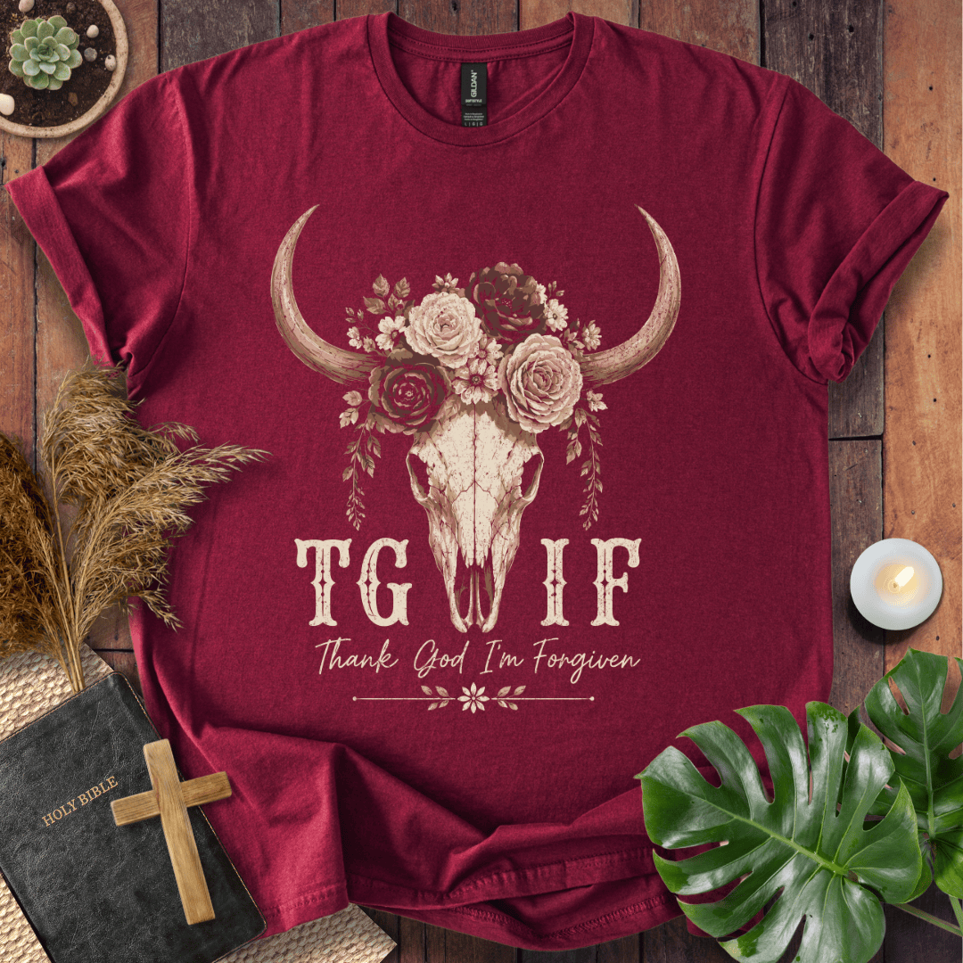 A maroon short-sleeve graphic T-shirt laid flat on a rustic wood surface, surrounded by decorative props including a Bible, cross, dried grass, monstera leaves, and a small candle. The shirt features a large central graphic of a skull with curved horns topped by flowers, with the text 'TG IF' and 'Thank God I'm Forgiven' printed below.