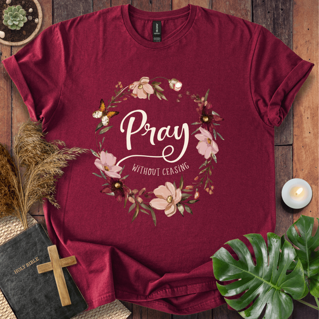 "Pray Without Ceasing" text inside a floral wreath with a butterfly on a shirt next to a Bible and cross.