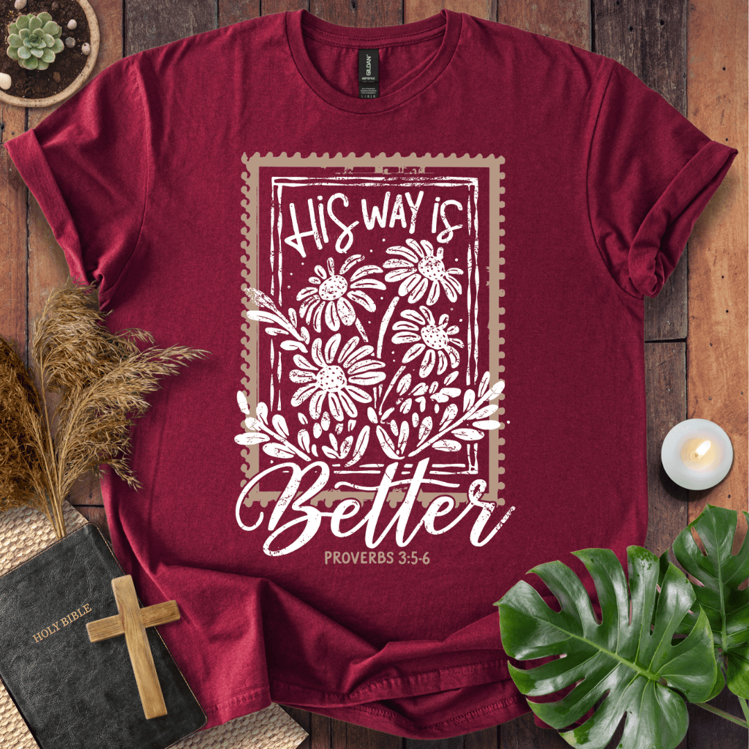 A maroon short-sleeve t-shirt with a white graphic print reading 'HIS WAY IS Better PROVERBS 3:5-6' with floral illustrations, styled in a flat-lay on a wooden surface with a book, cross, dried grass, succulent, and candle.