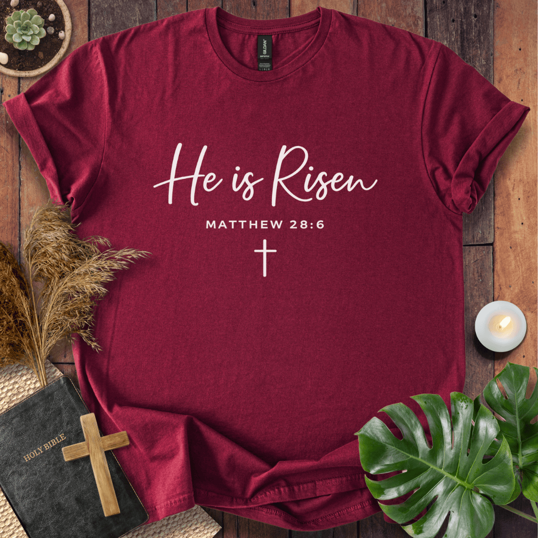 Easter Christian t-shirt with white "He is Risen" script and cross, styled on wood with a Bible and wooden cross.