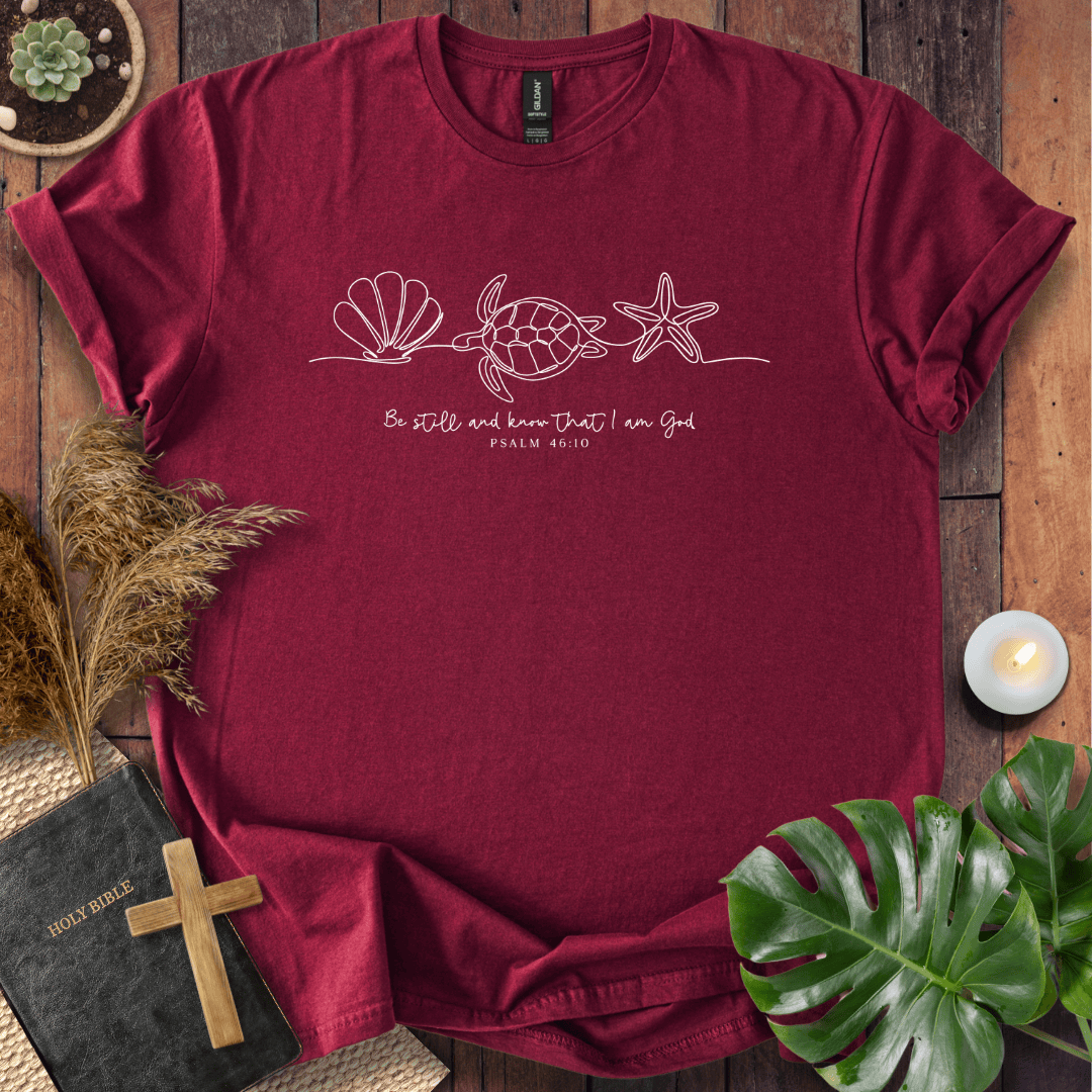 A maroon short-sleeve t-shirt with a white line art graphic of a sea turtle, starfish, and seashell, with the text 'Be still and know that I am God' and 'PSALM 46:10'. The shirt is displayed in a flat-lay arrangement on a wooden background with a bible, cross, dried grass, a plant, and a candle.