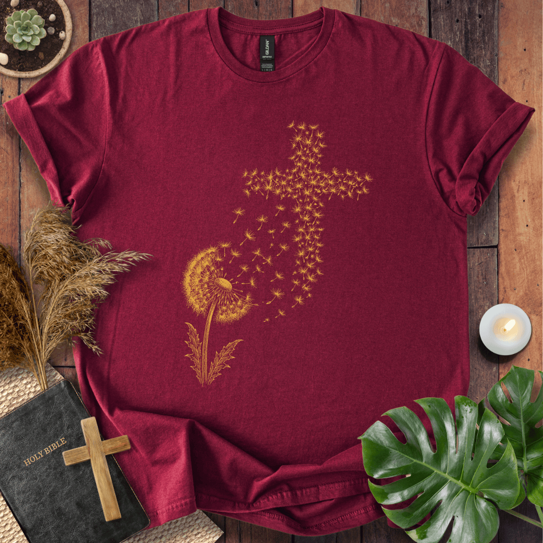 graphic t-shirt featuring a golden dandelion with seeds blowing in the wind to form a Christian cross shape.