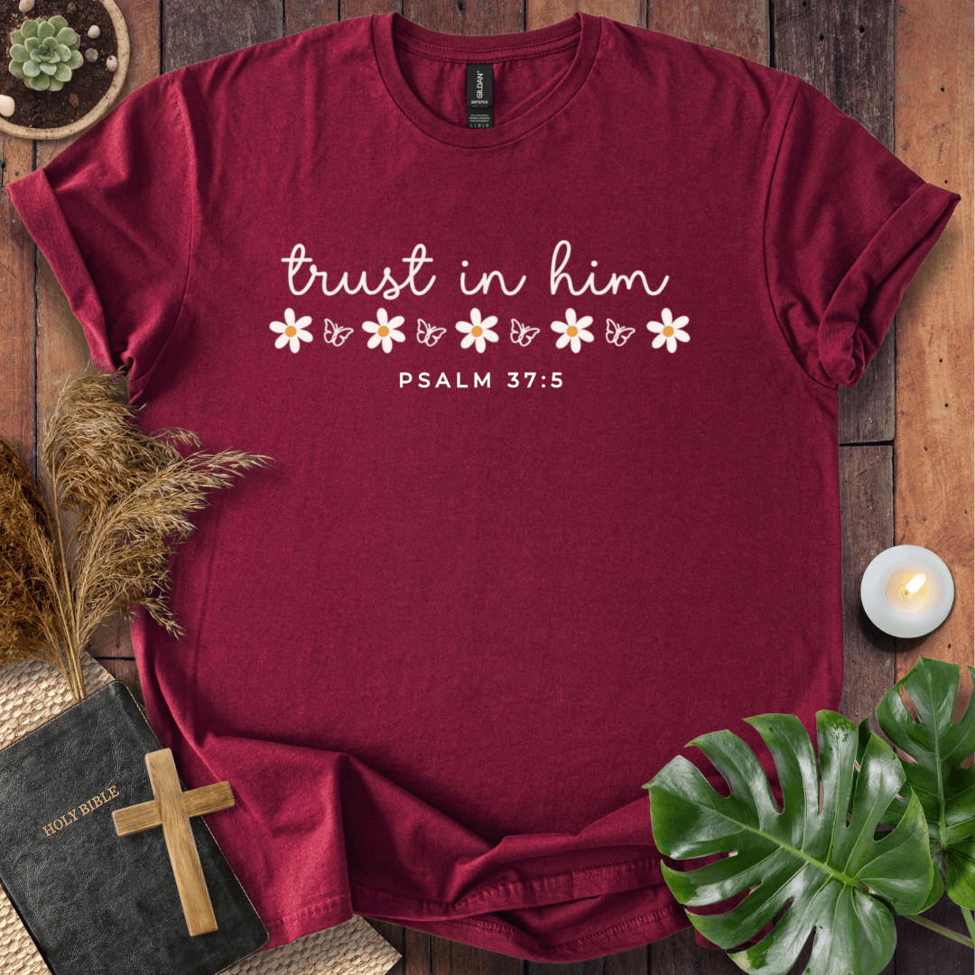 A maroon short-sleeve t-shirt with a white graphic print reading 'trust in him' and 'PSALM 37:5' with small flower illustrations, styled in a flat-lay on a wooden surface with a Bible, cross, plant leaves, and a candle.
