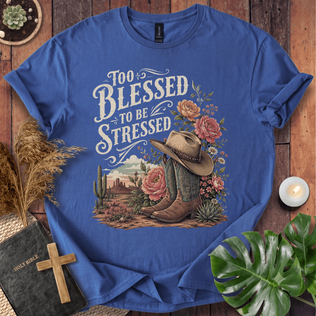 A blue graphic t-shirt laid flat on a rustic wooden surface, surrounded by decorative props including a Bible with a wooden cross, dried grass, a candle, and green leaves. The t-shirt features a western-themed graphic with the text 'Too Blessed to Be Stressed' and an illustration of cowboy boots, a cowboy hat, cacti, and flowers.