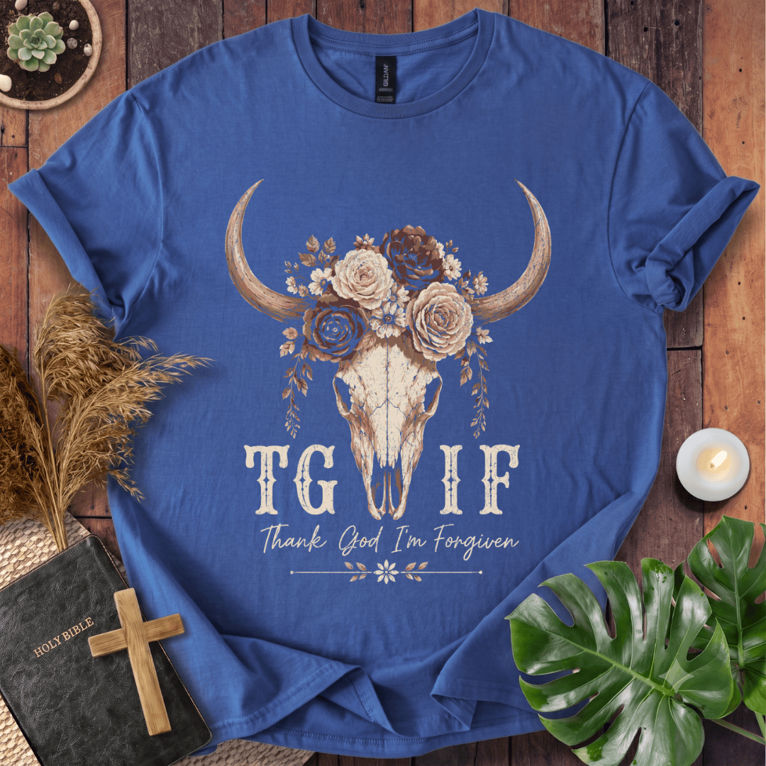 A royal blue short-sleeve graphic T-shirt laid flat on a rustic wooden surface, featuring a large floral and horned skull design above the text 'TG IF' and 'Thank God I'm Forgiven.' The shirt is styled with a Bible, cross, dried grass, candle, and green leaves around it.
