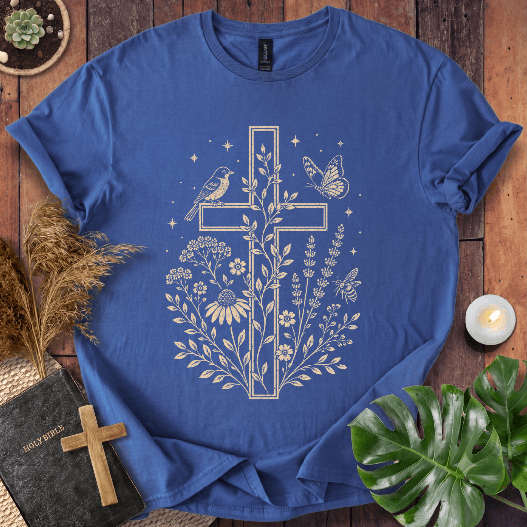 A blue short-sleeve crewneck t-shirt with a cream-colored graphic of a cross, flowers, birds, and butterflies, styled in a flat-lay on a wooden background with a bible, candle, and green leaves.