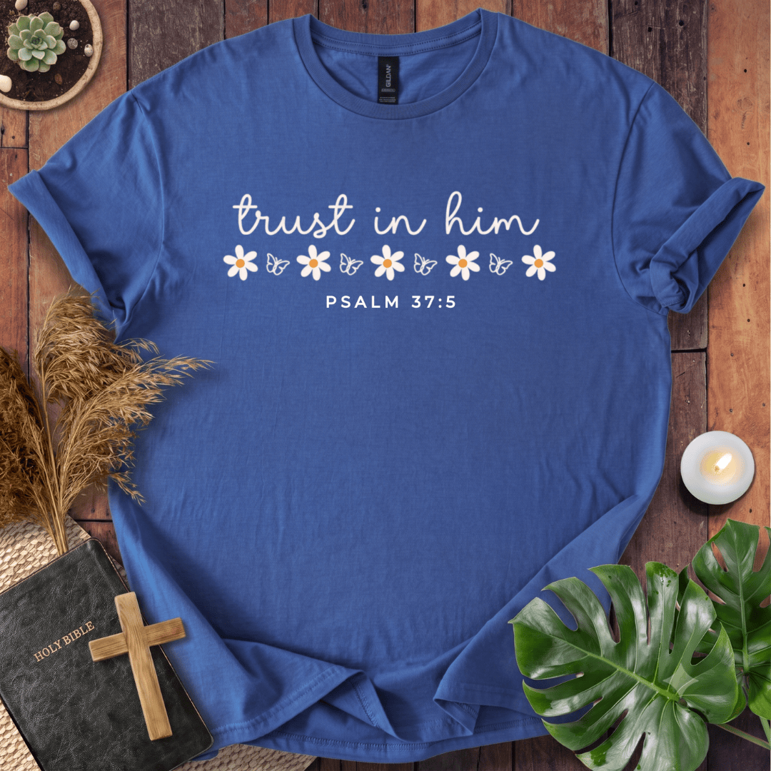 A blue short-sleeve T-shirt laid flat on a rustic wooden surface, printed with the phrase 'trust in him' in white script, a row of small white and yellow flowers, and 'PSALM 37:5' below. The shirt is styled with a book, cross, dried grass, candle, and green leaves around it.