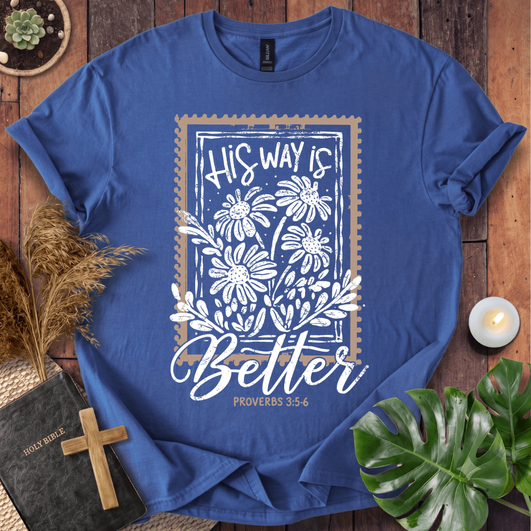 A blue graphic t-shirt laid flat on a rustic wooden surface, surrounded by decorative props including a small plant, dried grass, a Bible with a wooden cross, a candle, and a green monstera leaf. The shirt features a white floral design with the text 'His Way Is Better' and 'Proverbs 3:5-6'.