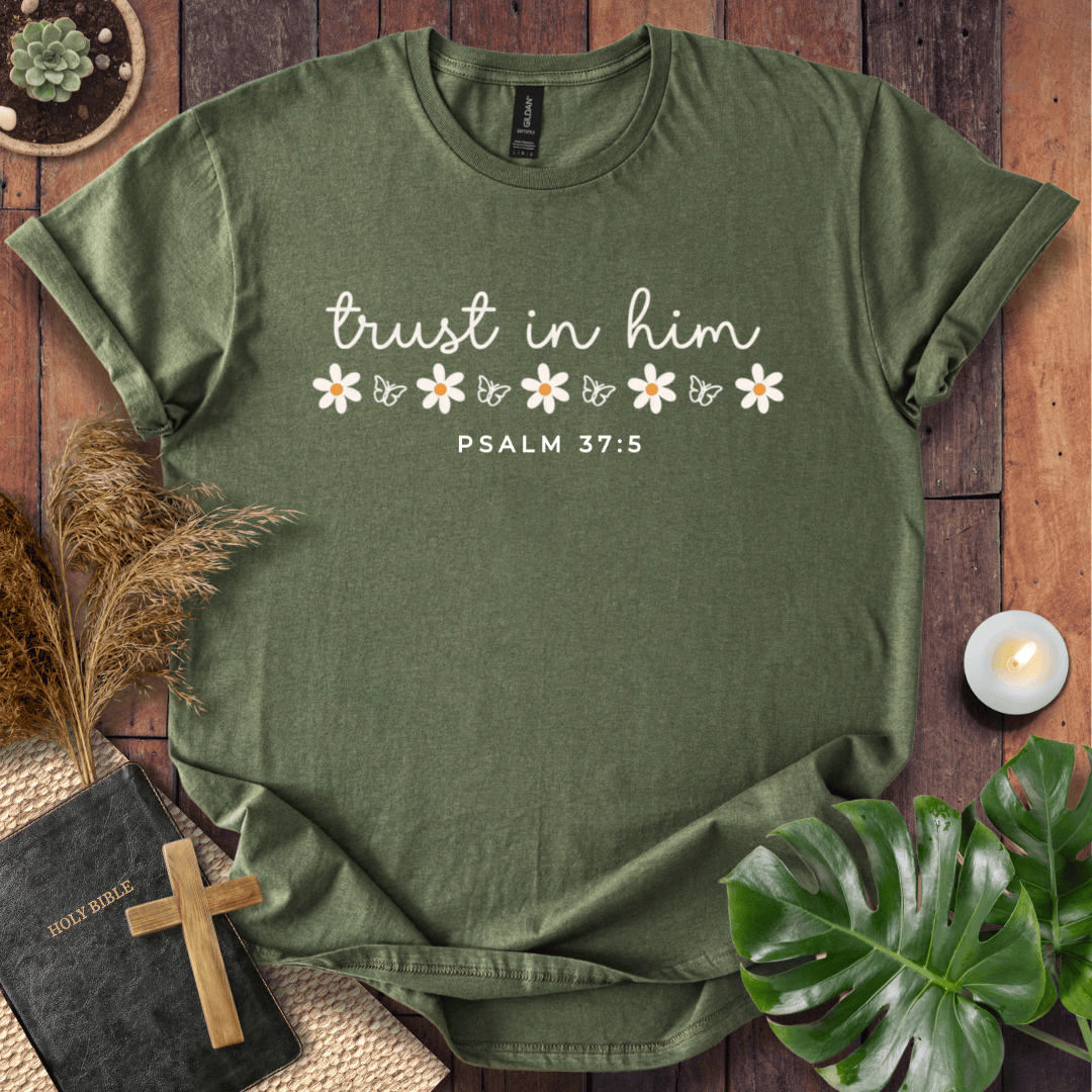 An olive green short-sleeve t-shirt with a white graphic print reading 'trust in him' and 'PSALM 37:5' with small daisy illustrations, laid flat on a rustic wooden background with decorative props including a Bible, cross, pampas grass, monstera leaf, and candle.