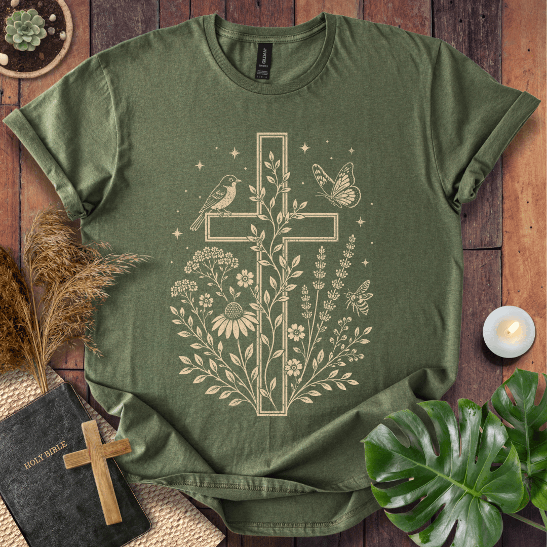 An olive green short-sleeve t-shirt with a beige graphic of a cross, flowers, birds, and a butterfly, presented in a flat-lay arrangement on a wooden surface with a Bible, candle, and green leaves.