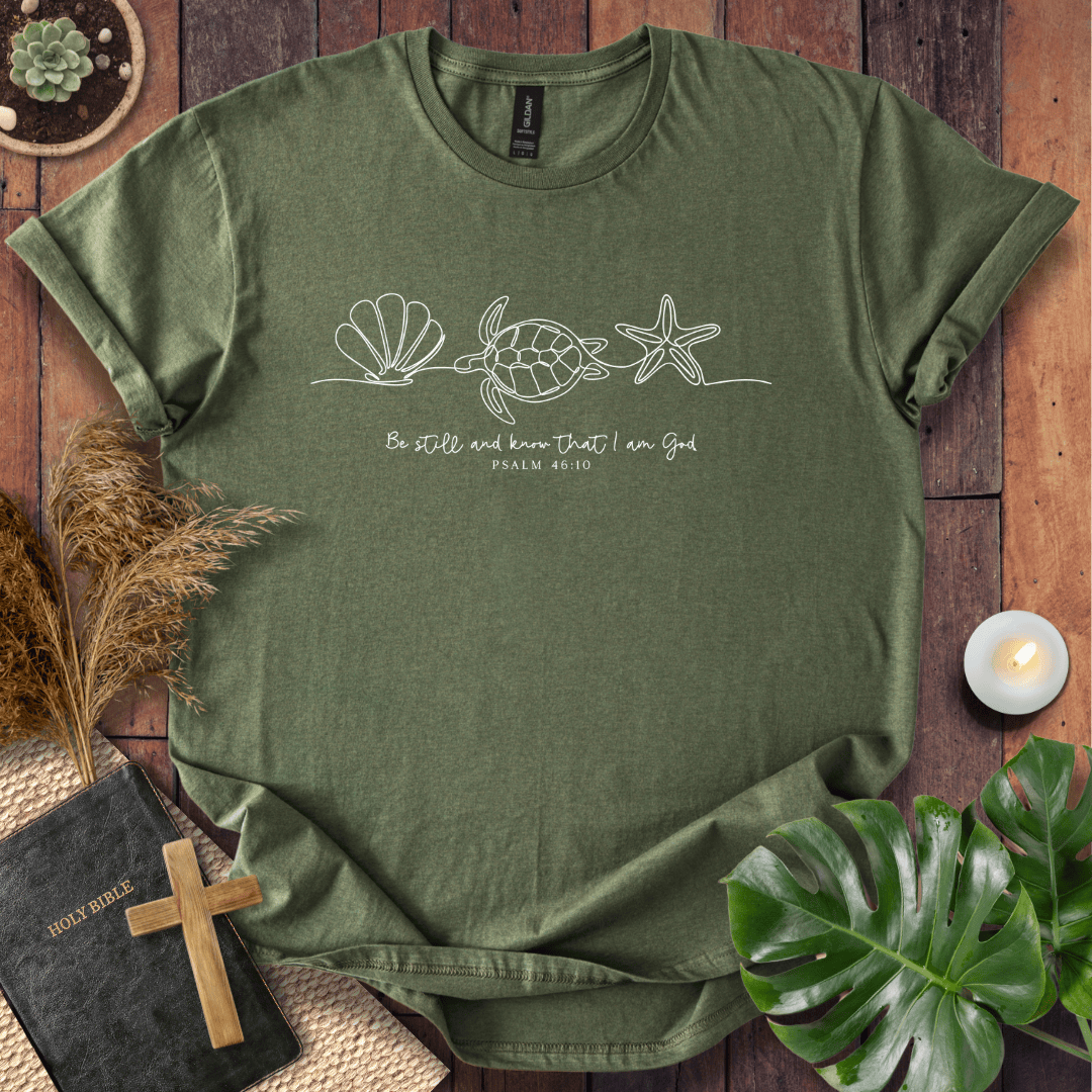 An olive green short-sleeve t-shirt with a white line art graphic of a sea turtle, seashell, and starfish, with the text 'Be still and know that I am God PSALM 46:10' printed on the chest. The shirt is displayed in a flat-lay arrangement on a rustic wooden background, surrounded by a bible, a wooden cross, pampas grass, a lit candle, and a green monstera leaf.
