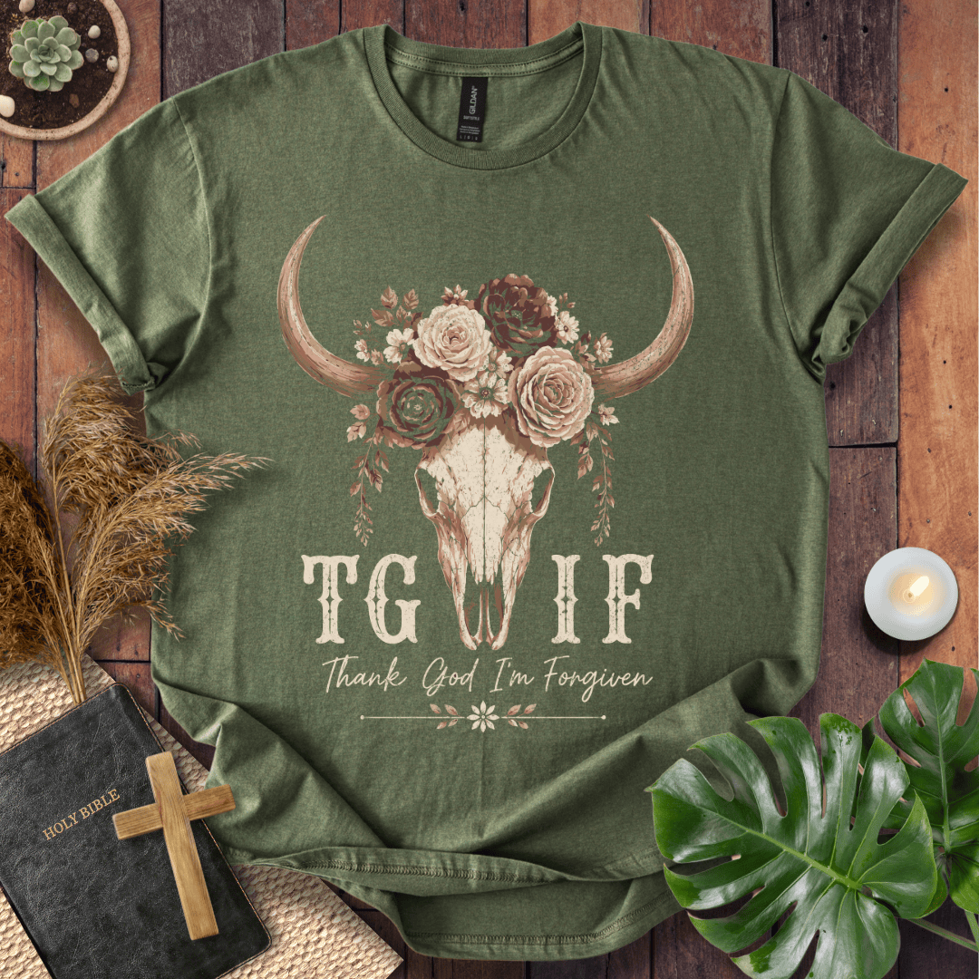 An olive green graphic t-shirt laid flat on a rustic wooden background with styled props including a bible, cross, dried grass, a candle, and a monstera leaf. The t-shirt features a central graphic of a skull decorated with a floral crown and the text 'TG IF Thank God I'm Forgiven'.