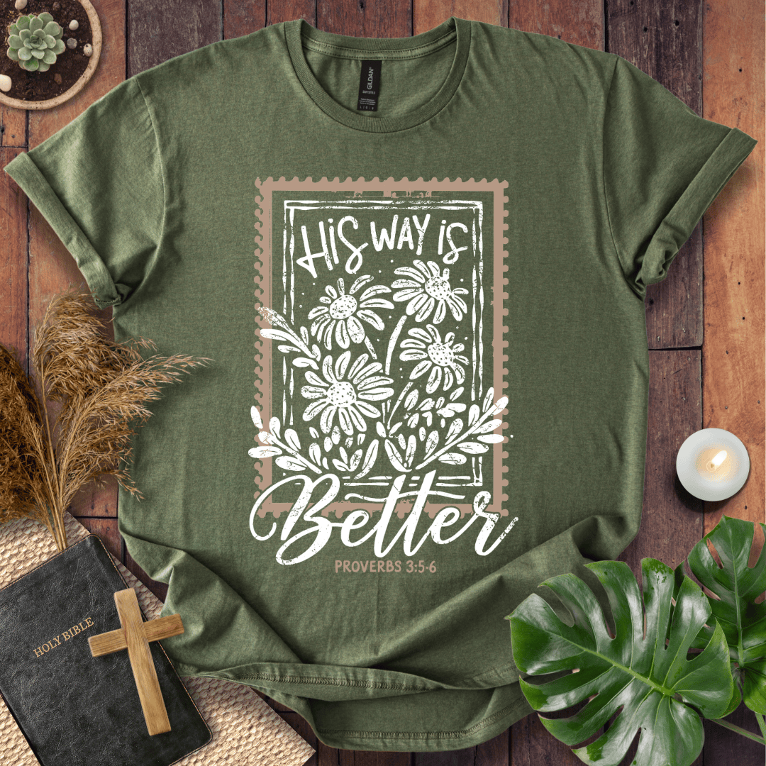 An olive green graphic t-shirt laid flat on a rustic wooden background, surrounded by decorative props including a small cross, a Bible, dried grass, a candle, and green leaves. The t-shirt features a white floral design with a brown border and the text 'HIS WAY IS Better PROVERBS 3:5-6'.