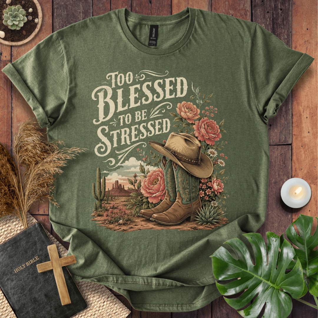 An olive green graphic t-shirt laid flat on a rustic wooden background, surrounded by decorative props including a Bible, a wooden cross, pampas grass, a lit candle, and a green monstera leaf. The t-shirt features a western-themed graphic with the text 'Too Blessed to Be Stressed' above an illustration of a cowboy hat, boots, cactus, and pink roses.