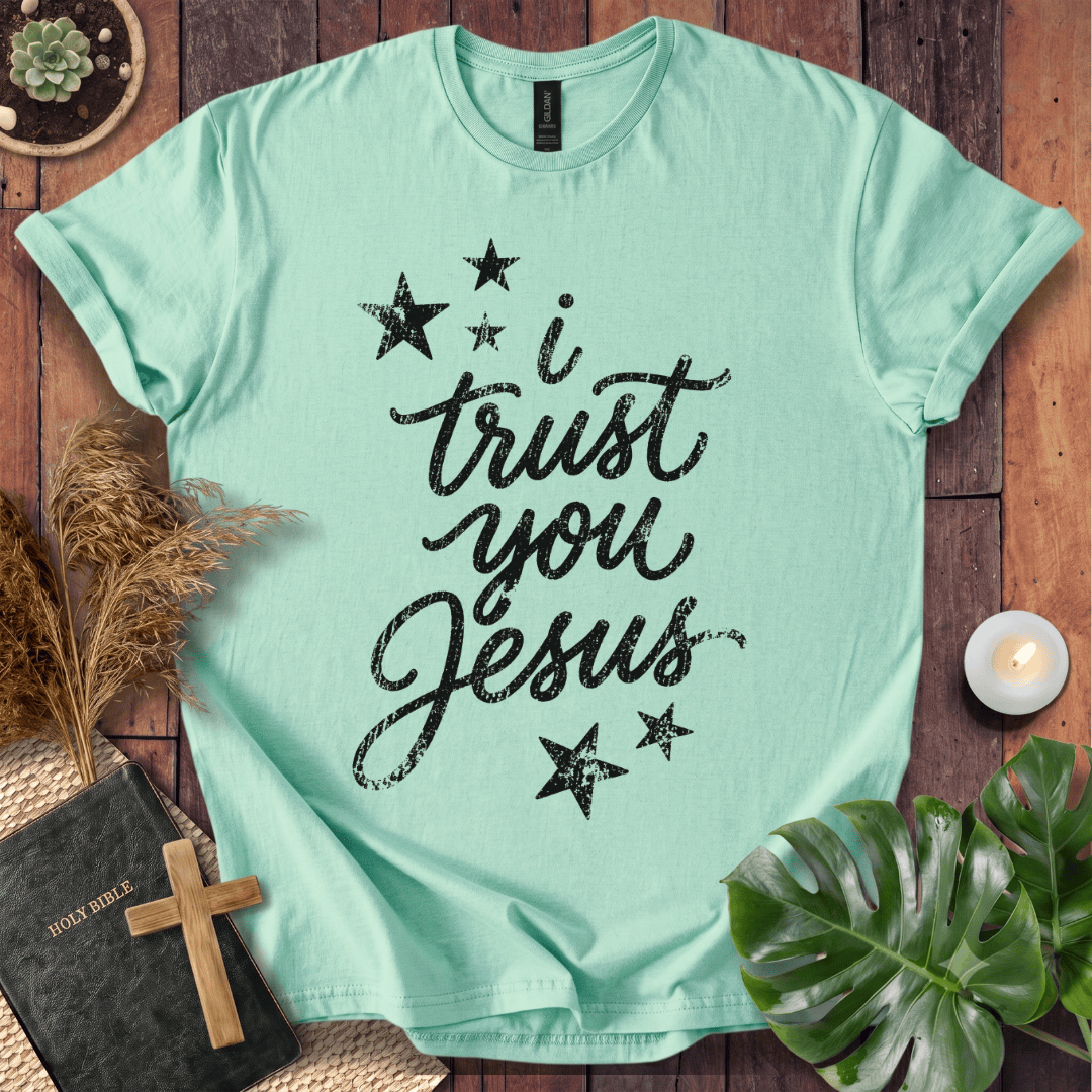A mint green short-sleeve t-shirt with the text 'i trust you Jesus' in black brush script and black stars, presented in a flat-lay arrangement on a wooden background with a bible, cross, pampas grass, a plant, and a candle.