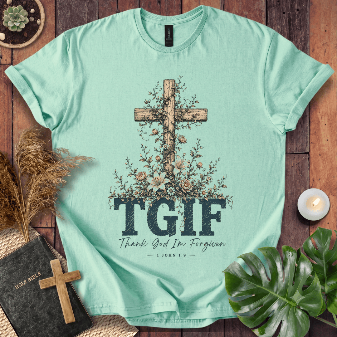 A mint green graphic t-shirt laid flat on a rustic wooden background, surrounded by decorative props including a Bible, a small wooden cross, dried grass, a lit candle, and green leaves. The t-shirt features a large wooden cross decorated with flowers, the text 'TGIF' in dark blue block letters, and 'Thank God I'm Forgiven — 1 JOHN 8:32' underneath.