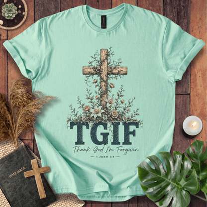 A mint green graphic t-shirt laid flat on a rustic wooden background, surrounded by decorative props including a Bible, a small wooden cross, dried grass, a lit candle, and green leaves. The t-shirt features a large wooden cross decorated with flowers, the text 'TGIF' in dark blue block letters, and 'Thank God I'm Forgiven — 1 JOHN 8:32' underneath.