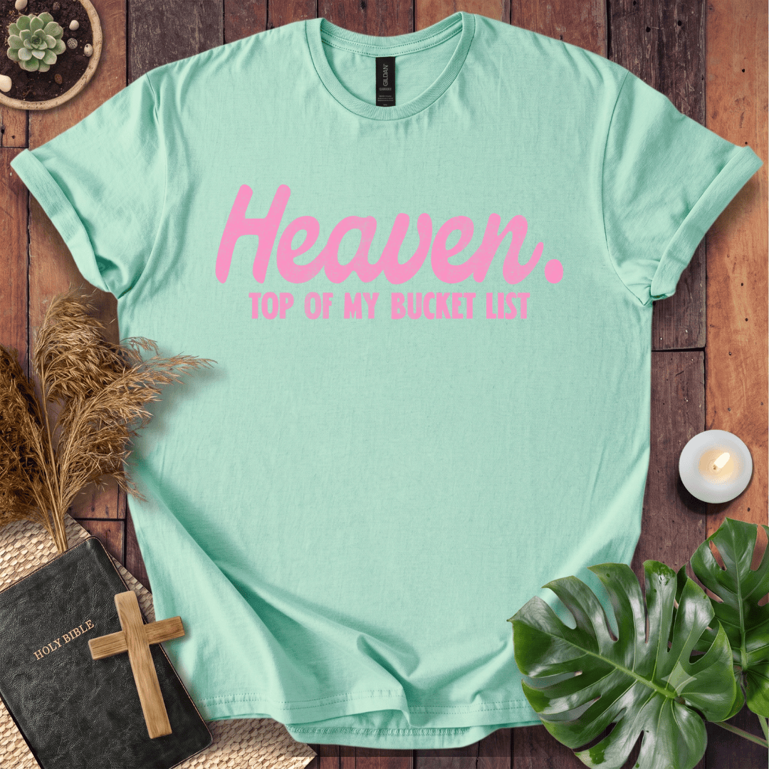 A mint green short-sleeve t-shirt with pink text reading 'Heaven. TOP OF MY BUCKET LIST' is laid flat on a rustic wooden surface with decorative props including a book, cross, dried grass, candle, plant leaves, and a small pot.