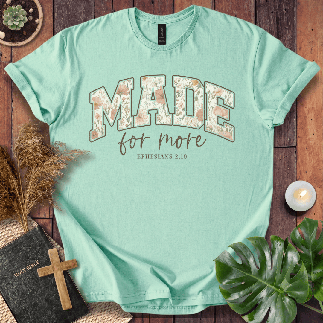 A mint green short-sleeve t-shirt with a floral-patterned 'MADE' graphic and the text 'for more EPHESIANS 2:10' printed on the front. The shirt is displayed in a flat-lay arrangement on a rustic wooden background, surrounded by a holy book with a wooden cross, pampas grass, a small candle, and a green monstera leaf.