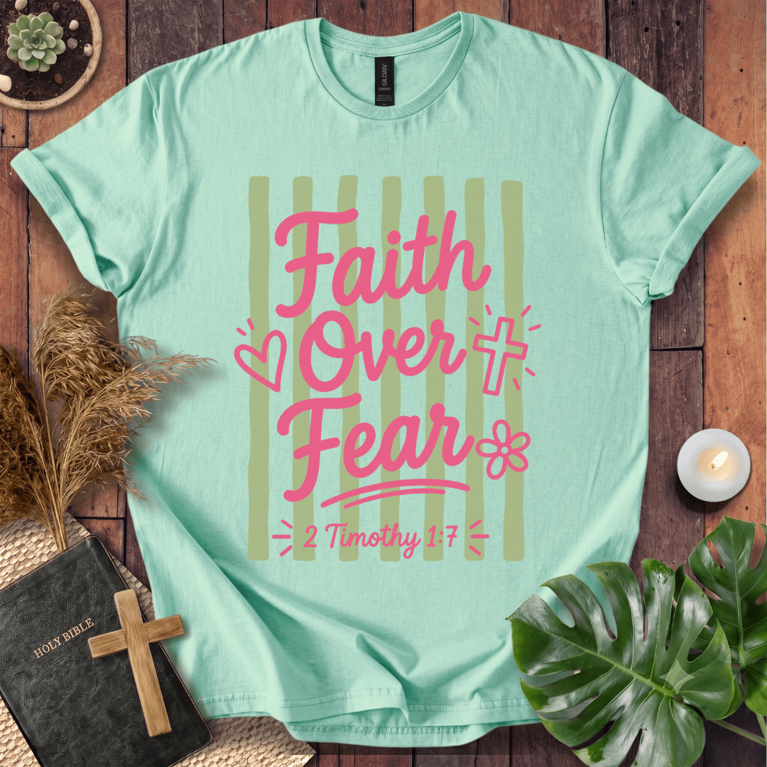 A mint green graphic t-shirt laid flat on a rustic wooden background, surrounded by decorative props including a Bible with a wooden cross, dried grass, a small plant, a candle, and a monstera leaf. The t-shirt features a graphic with the text 'Faith Over Fear' and '2 Timothy 1:7' in pink lettering over light olive-green vertical stripes.