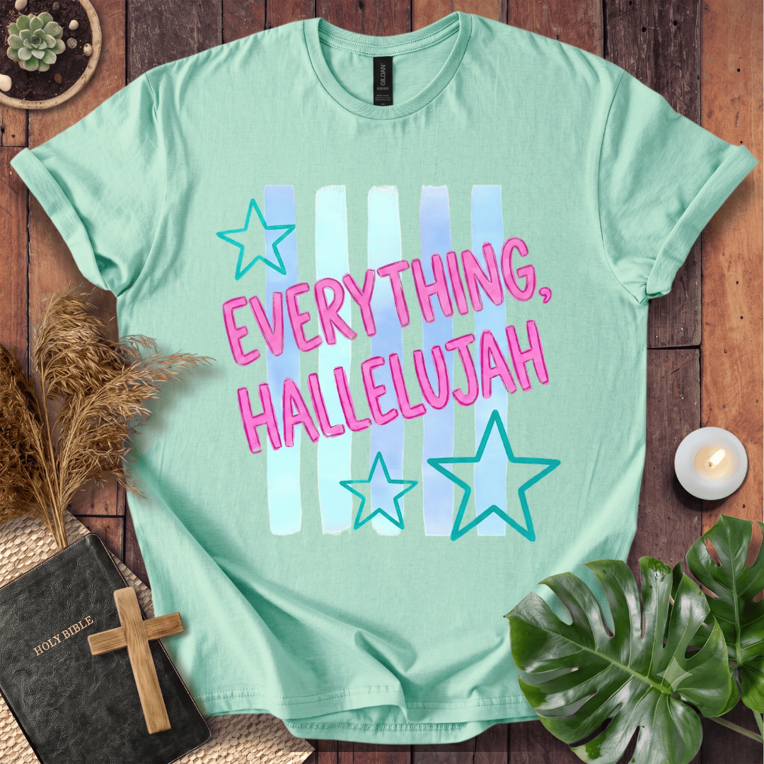 A mint green graphic t-shirt laid flat on a rustic wooden background, surrounded by decorative props including a small plant, a candle, a book with a wooden cross, and green leaves. The t-shirt features the text 'EVERYTHING, HALLELUJAH' in pink letters with light blue and white vertical stripes behind it, accented by two outlined teal stars.